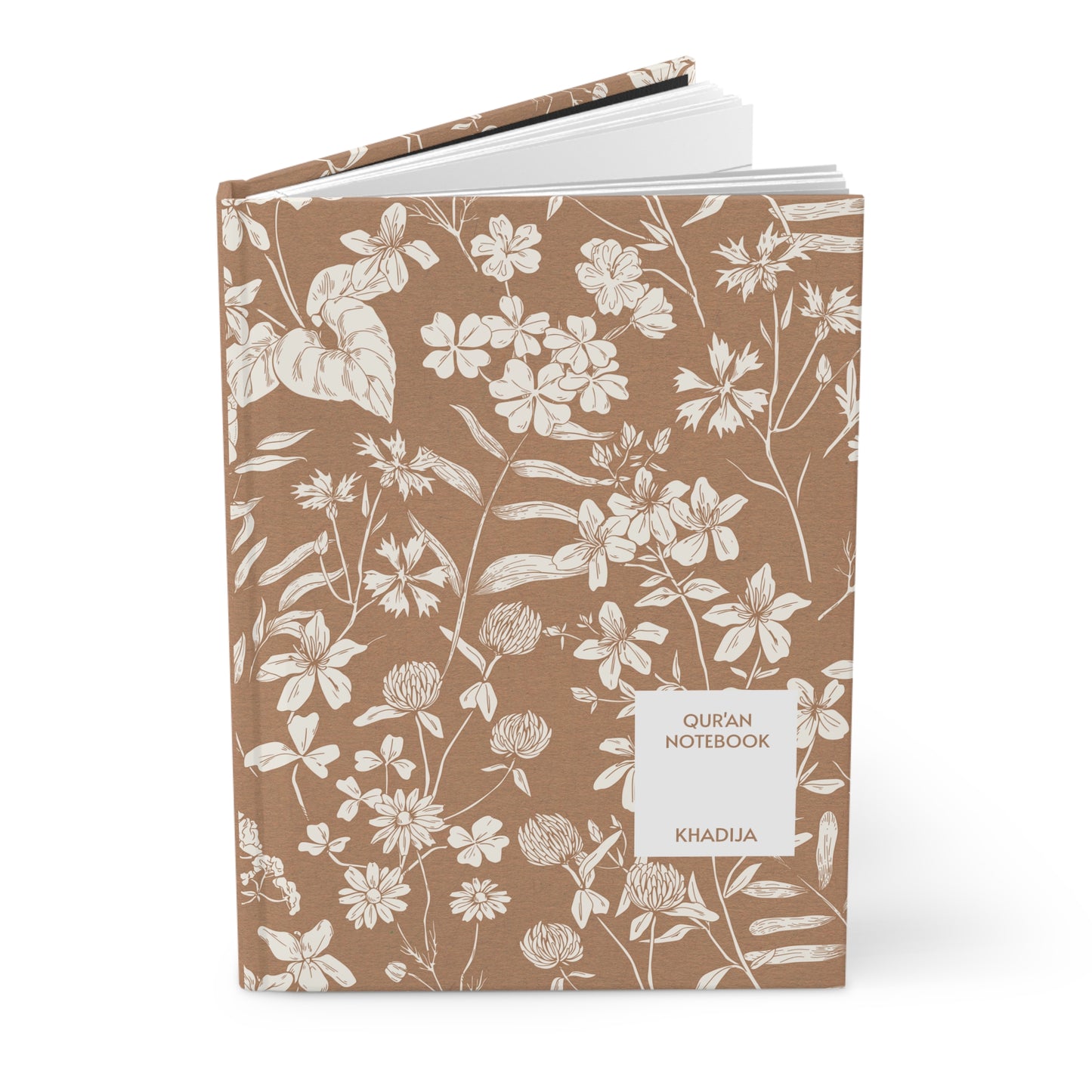 Angled view of a Quran notebook with white floral art on a brown background.